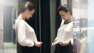 Closeup of attractive brunette standing in front of a big mirror at retail store and trying on a white blouse and black skirt. She seems happy with what she sees. Side view, selective focus.