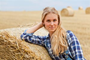 Beautiful young woman standing on field and leaning on roll bale of straw. She contentedly looking into the camera. Shallow DOF. Shooting with the Canon EOS 5D Mark II.